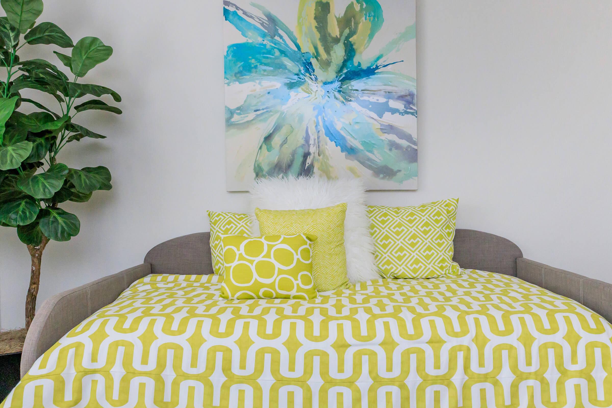 A cozy seating area featuring a gray couch with a patterned yellow and white blanket. Several decorative pillows in varying patterns and shades of yellow sit on the couch. Above it hangs a colorful floral artwork. A large leafy plant is positioned to the left.