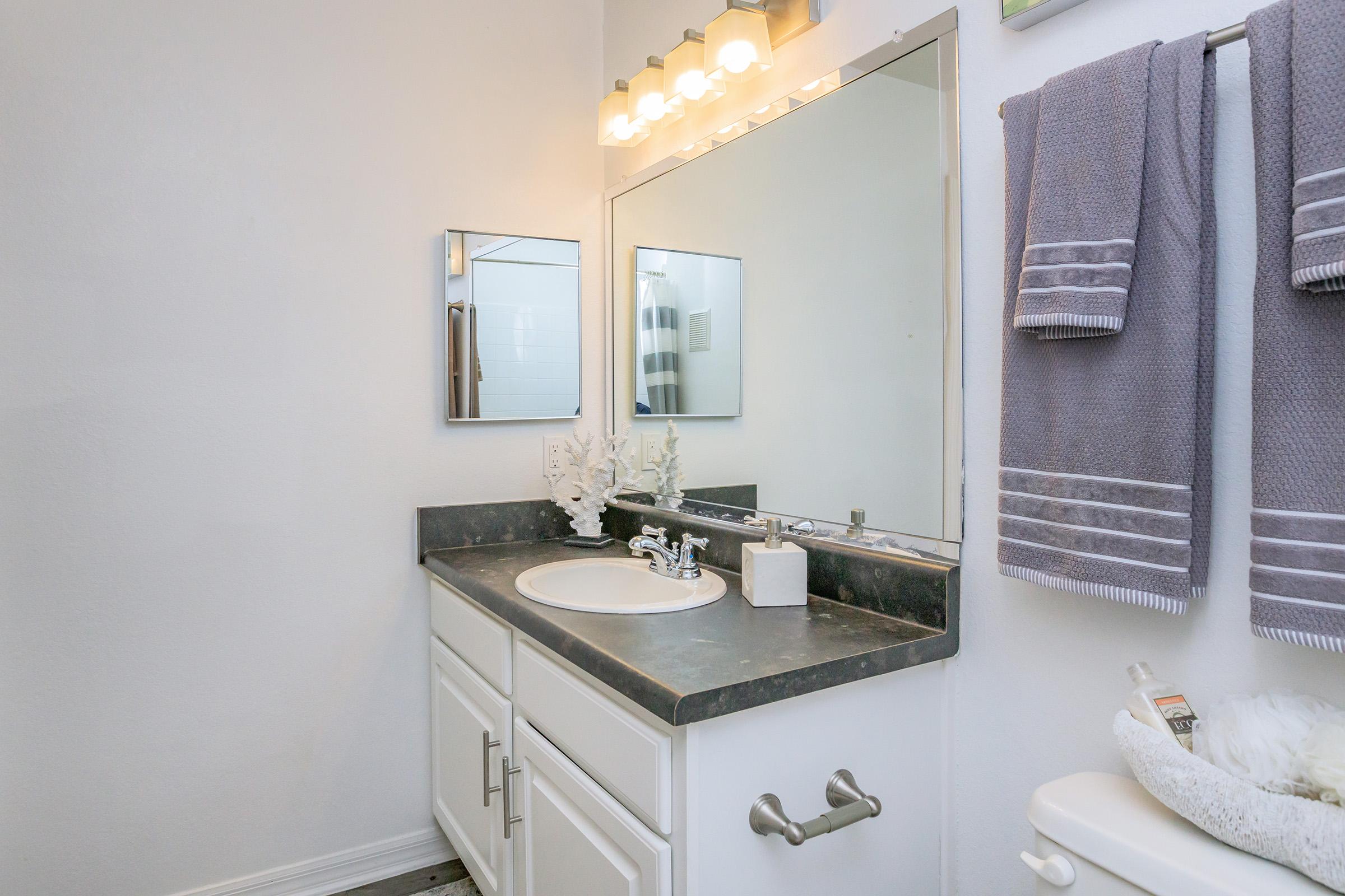 A clean and modern bathroom featuring a white vanity with a round sink, a large mirror above it, and a light fixture with three bulbs. There are gray towels hanging on a towel rack, and decorative items on the countertop. The walls are painted white, and the flooring appears to be a light wood.