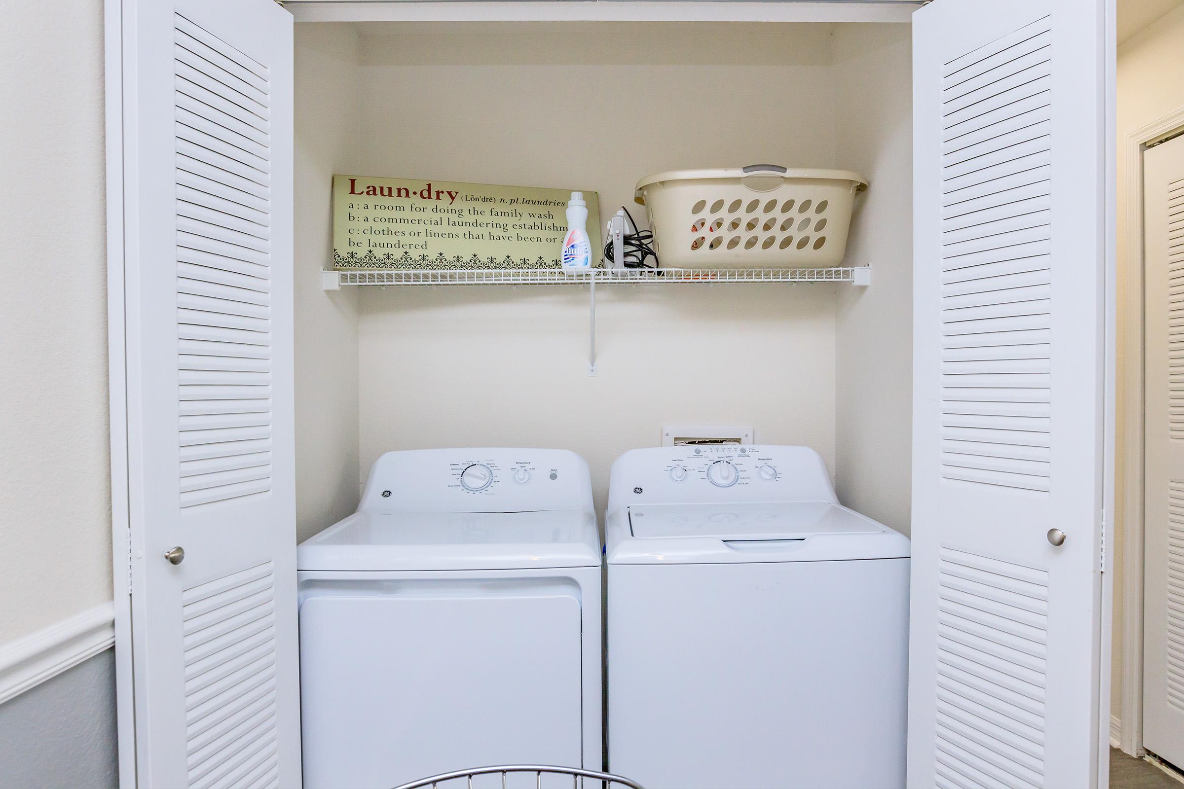 A small laundry area with two white washing machines side by side, located behind folding doors. Above them, a shelf holds a laundry basket and a spray bottle. A decorative sign on the wall reads about laundry as an essential household task. The overall space is tidy and organized.