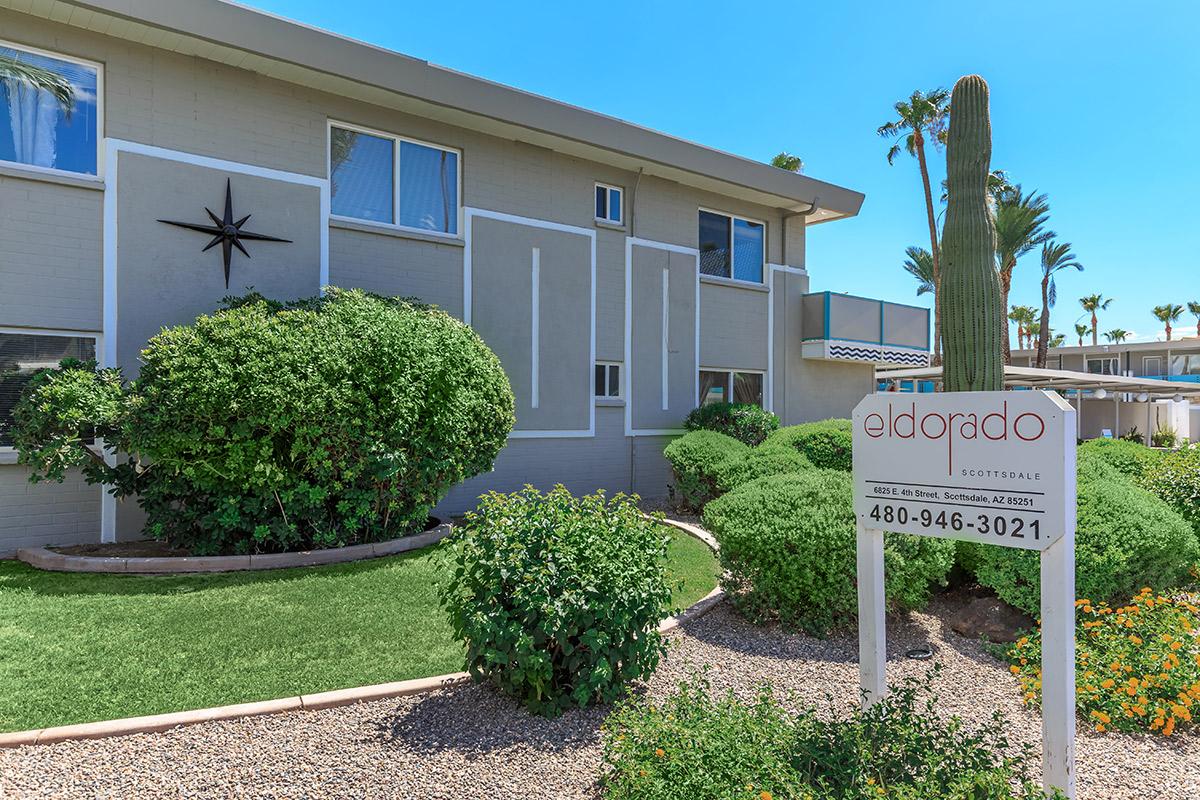 A well-maintained apartment building with a landscaped front yard featuring shrubs and a tall cactus. A sign indicating "Eldorado" is prominently displayed in front, along with contact information. The sky is clear and blue.