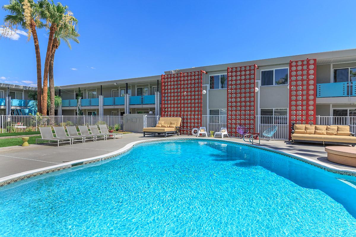 A sunny outdoor pool surrounded by lounge chairs, with a backdrop of a modern multi-story building featuring red and white patterned walls and palm trees.