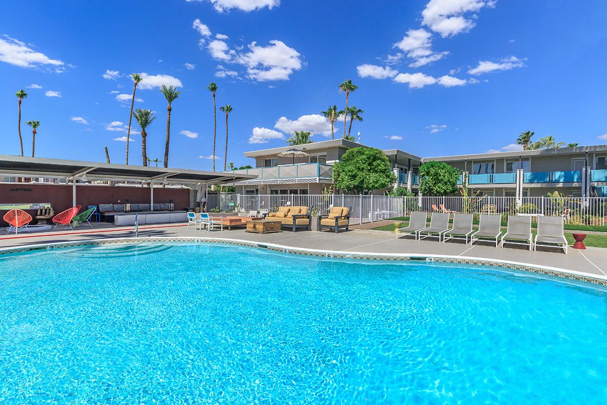 A clear blue swimming pool surrounded by lounge chairs and palm trees, with an apartment building in the background under a bright blue sky with scattered clouds.