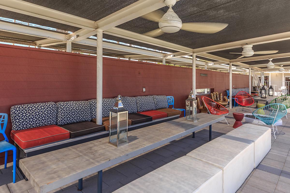 A spacious outdoor seating area with colorful furniture, including patterned couches and red and blue chairs. There is a long wooden table in the center, and the area is shaded by a canopy with ceiling fans. The walls are painted in a warm color, enhancing the inviting atmosphere.