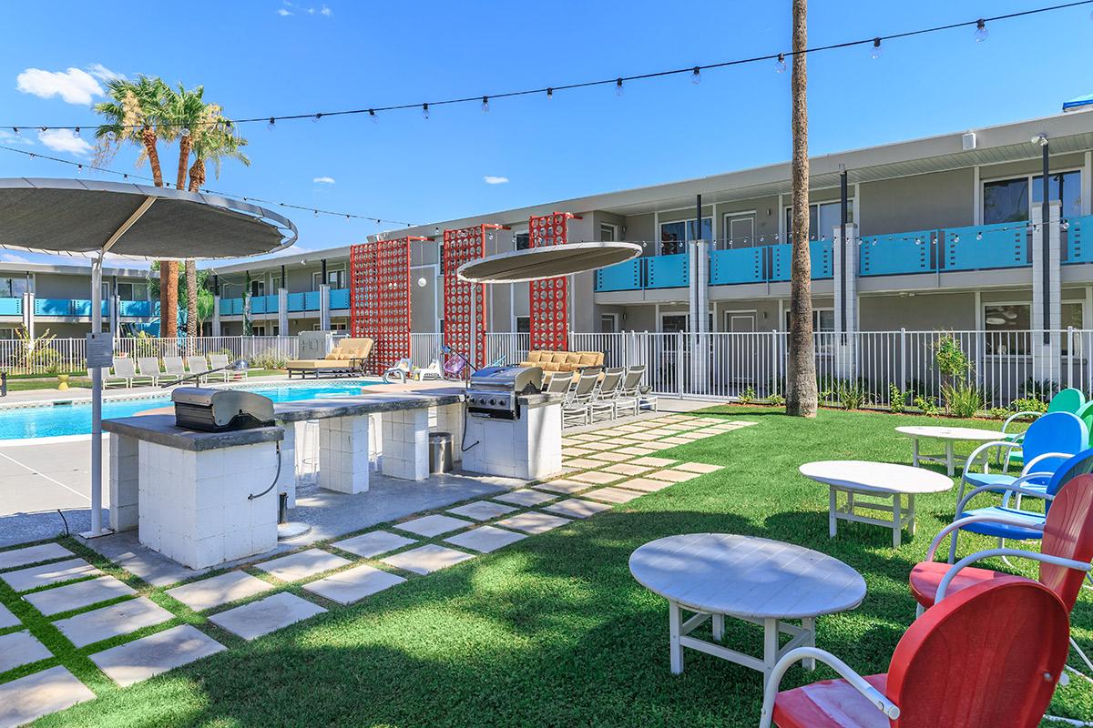 A bright pool area with lounge chairs, outdoor seating, and a barbecue grill, surrounded by palm trees and a modern hotel building in the background. The sky is clear and blue, with string lights overhead.