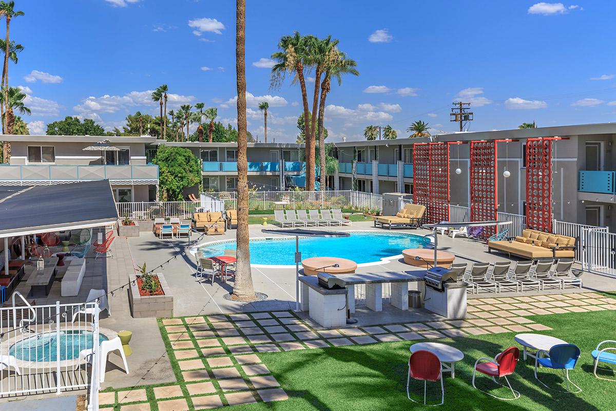 A sunny outdoor pool area surrounded by palm trees and lounge chairs, with a modern building in the background. Colorful patio furniture is arranged around the pool, creating a vibrant atmosphere.