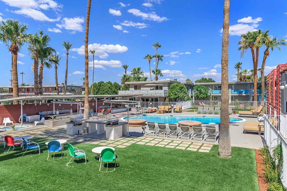 A bright outdoor pool area surrounded by palm trees, featuring lounge chairs, green grass, and a barbecue grill. The sky is clear with a few clouds, creating a vibrant and inviting atmosphere.