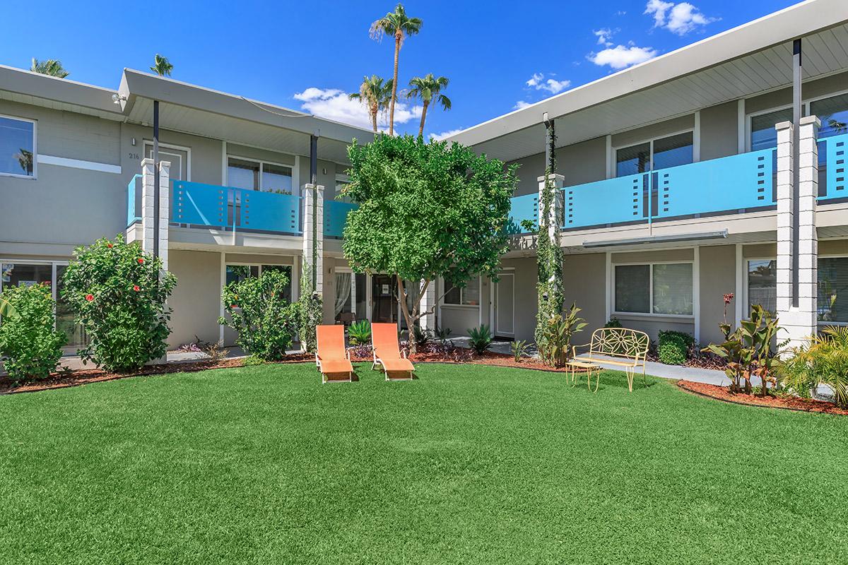 A well-maintained courtyard of an apartment community featuring two lounge chairs on a grassy area, surrounded by various plants and trees, with balconies visible in the background under a blue sky.