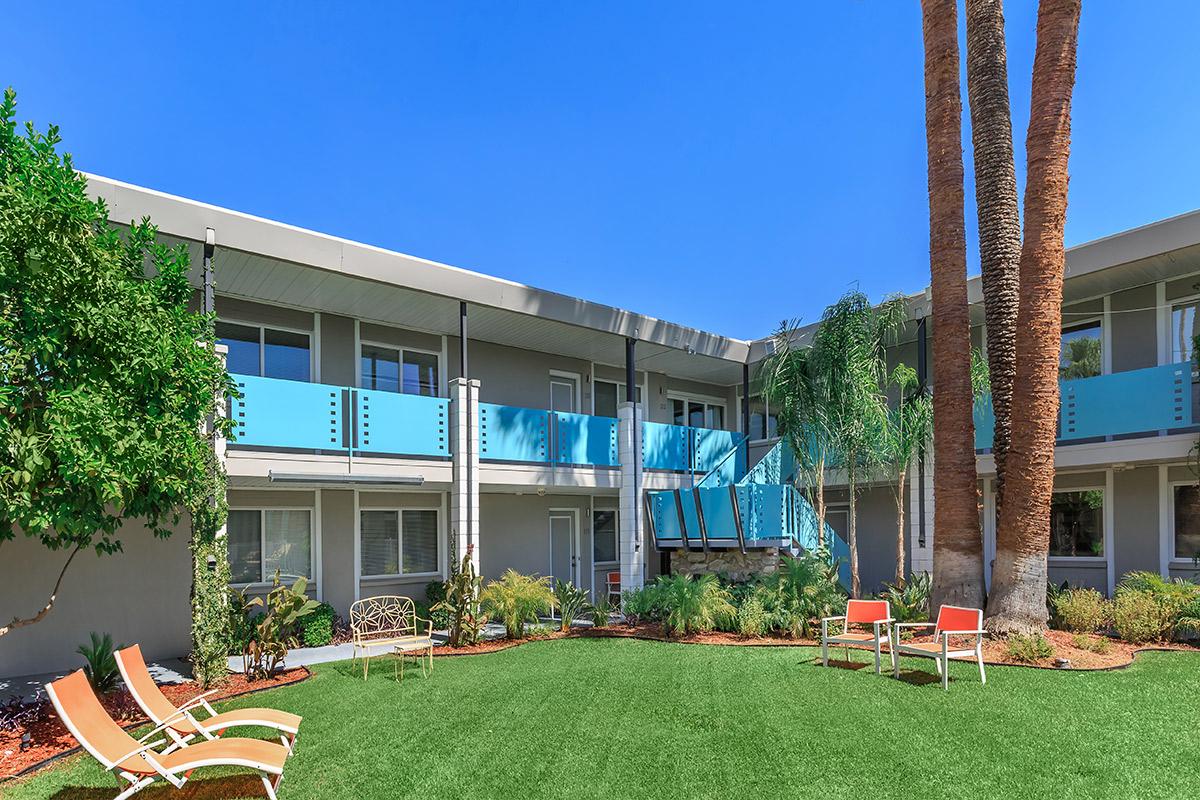 A sunny courtyard surrounded by a two-story building with balconies. The area features green grass, palm trees, and seating options, including lounge chairs and benches.