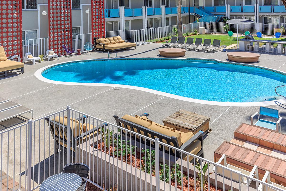 A swimming pool surrounded by lounge chairs and tables, with a colorful building in the background and well-maintained landscaping.