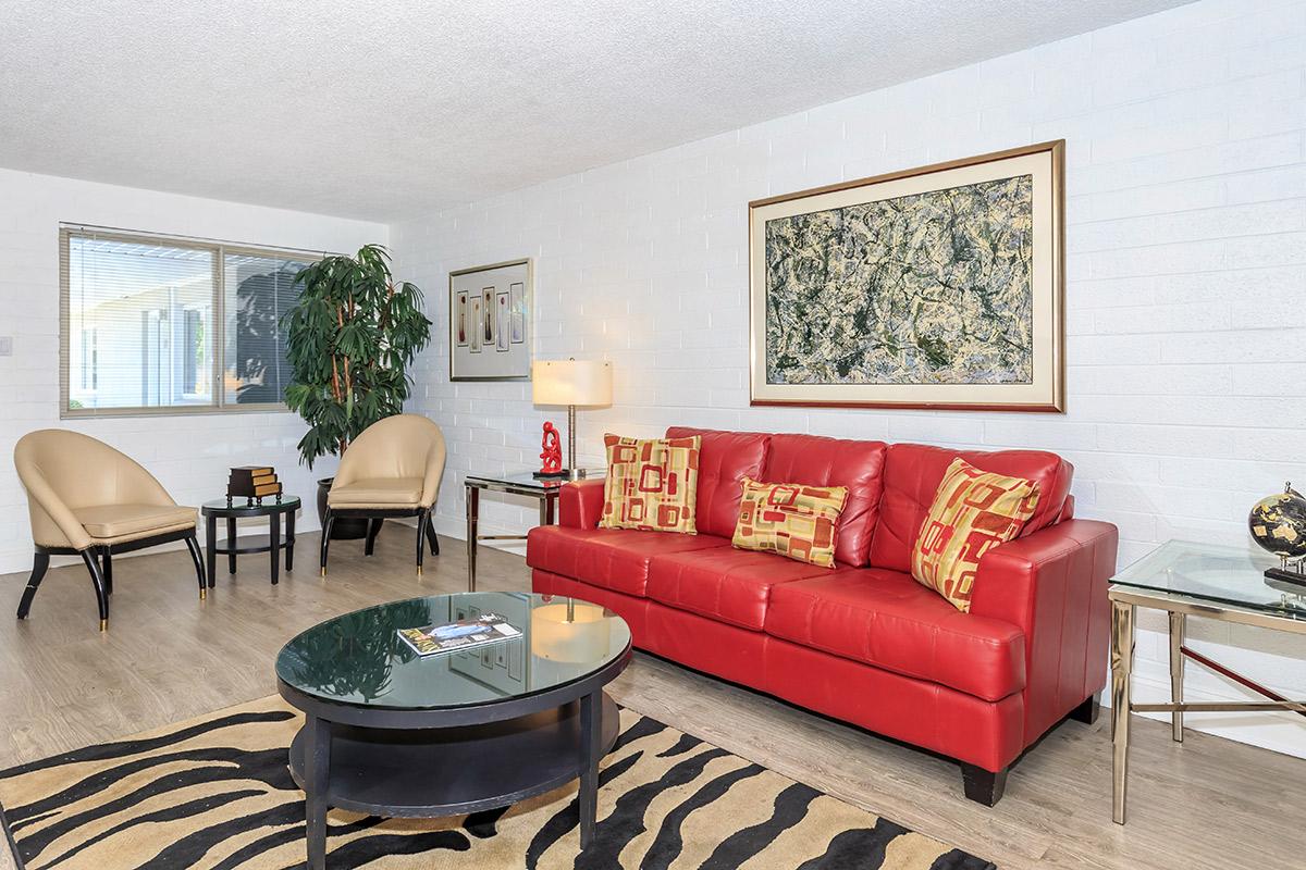A living room featuring a red leather sofa with decorative pillows, two beige armchairs, a glass coffee table on a tiger-striped rug, and a large piece of artwork on the wall. There is a potted plant in the corner and a lamp beside the sofa.