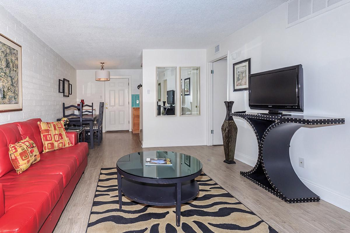 A modern living room featuring a red couch with decorative pillows, a round black coffee table, and a zebra-striped area rug. In the background, there is a dining area with a light fixture, a mirrored wall, and a television on a stylish console. The walls are painted white, and there are framed artworks and a decorative vase.