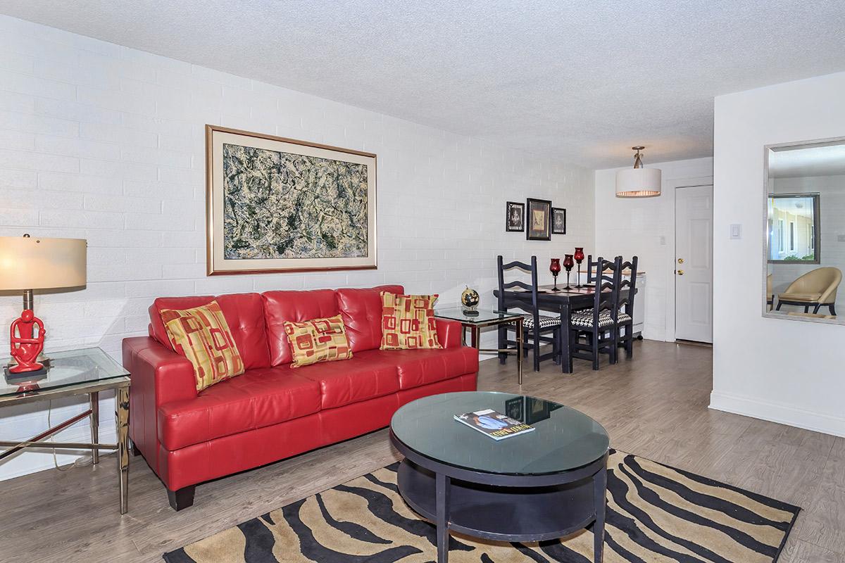 A modern living room featuring a red leather sofa with decorative pillows, a glass coffee table, and a zebra-striped rug. There is a dining area with a dark wooden table and black chairs adorned with red candles, and a wall-mounted mirror reflects the space. Light-colored walls and a piece of artwork add to the decor.