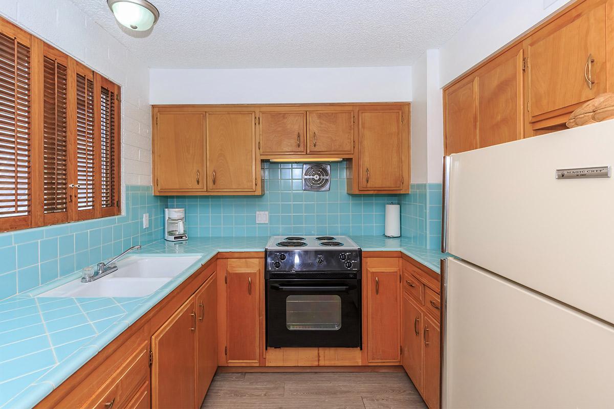 A cozy kitchen featuring wooden cabinets, a light blue tiled backsplash, a stovetop, and a white refrigerator. The kitchen has a window with wooden blinds, providing natural light.