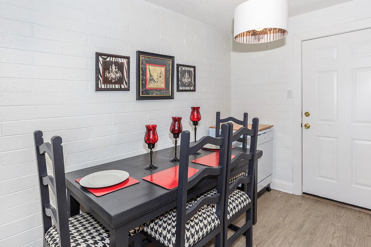 A modern dining area featuring a dark wooden table with four chairs, each with checkered cushions. The table is set with red decorative plates and surrounded by red candle holders. Two framed artworks are displayed on the white brick wall. A light fixture hangs above the table, and a door leads out of the room. The flooring is a light wood finish.
