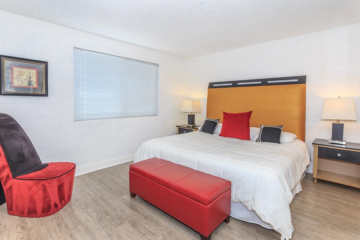 A modern bedroom featuring a large bed with a yellow headboard, white bedding, and decorative pillows. There are two bedside lamps on either side, a red bench at the foot of the bed, and a stylish red accent chair. A window allows natural light to flow in, and the flooring is a light wooden texture.