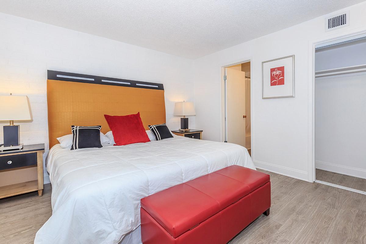 A modern bedroom featuring a large bed with a yellow headboard and red accent pillows. There are two bedside lamps on nightstands, and a red bench at the foot of the bed. A closet is visible on the right side, with a door leading to another room. The walls are painted white and the flooring is light wood.