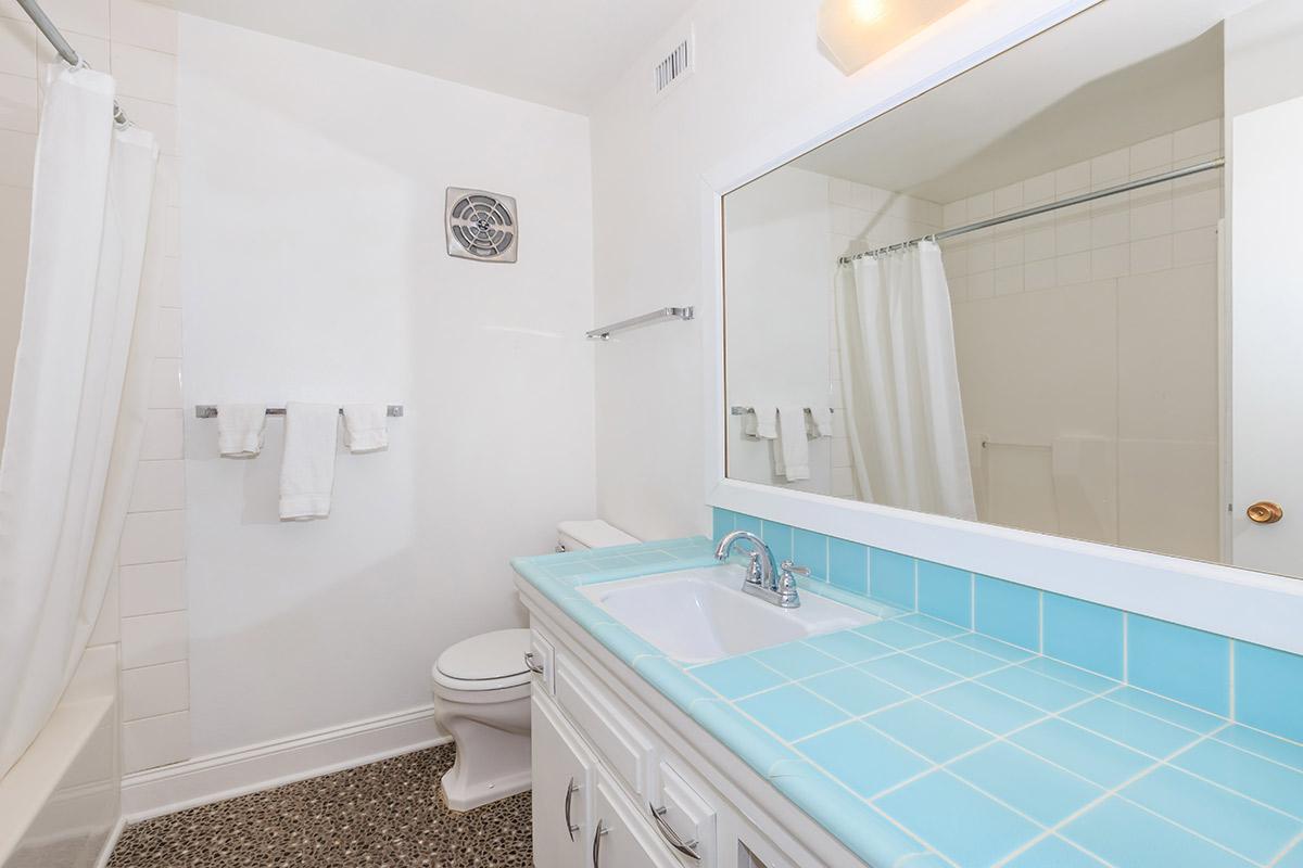 A clean, modern bathroom featuring a white bathtub, a toilet, and a large mirror above a light blue countertop with a sink. Two rolled white towels hang on a rack, and there is a small ventilation fan in the ceiling. The walls are painted white, and the floor has a textured design.