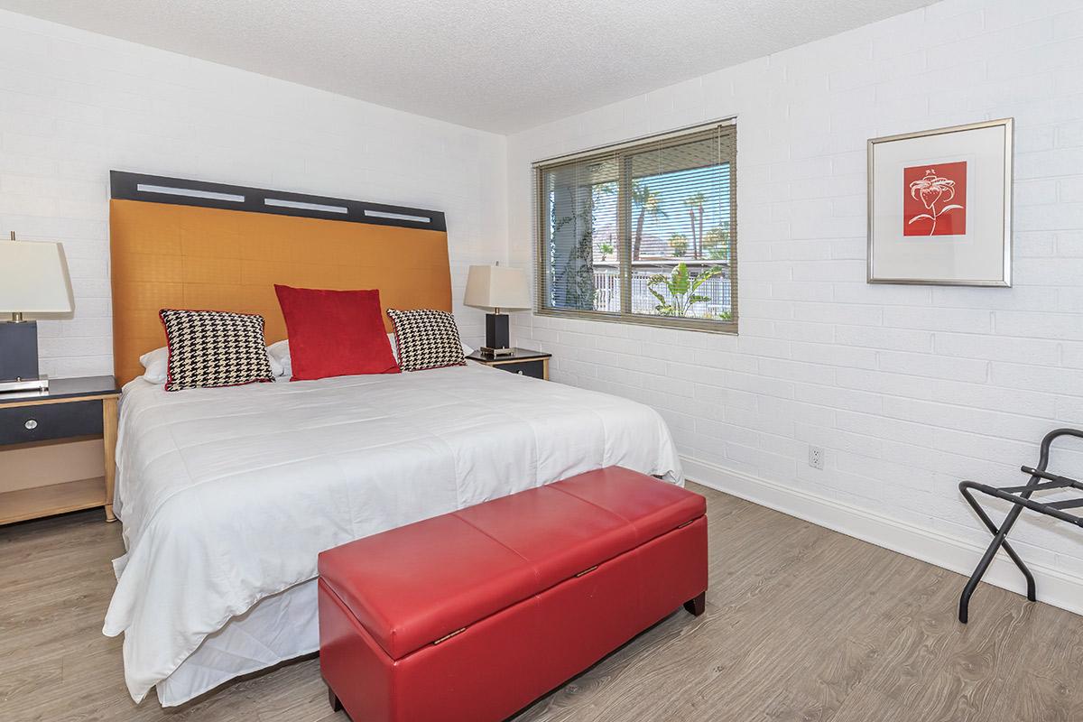 A modern bedroom featuring a queen-sized bed with white bedding and colorful accent pillows. There are two bedside tables with lamps, a red bench at the foot of the bed, and a window with blinds allowing natural light. The floor has wood-like tiles, and a piece of artwork is hung on the wall.