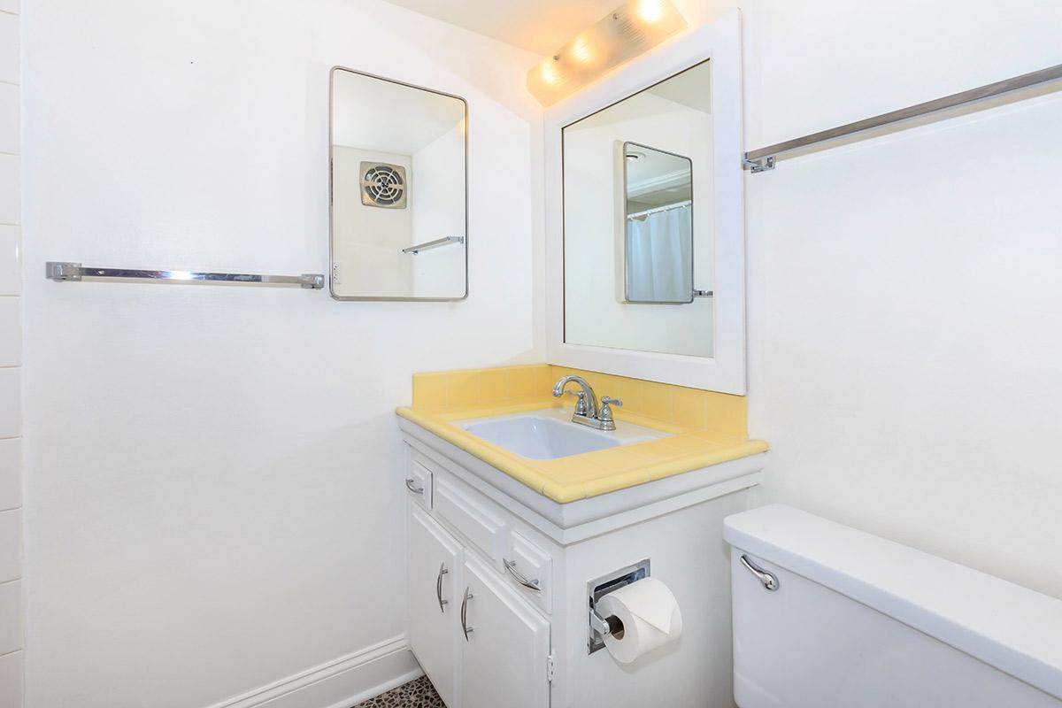 A clean, modern bathroom featuring a yellow countertop sink, a wall-mounted mirror, a toilet, and a towel rack. The walls are painted white, with simple decor and tiled flooring.