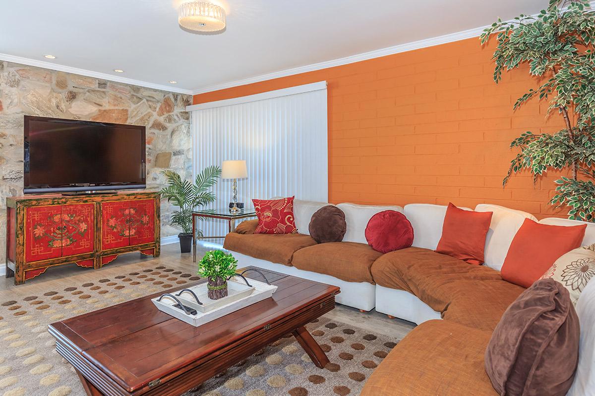 A cozy living room featuring a large sectional sofa with brown and white cushions, a wooden coffee table with a decorative tray and a small plant, a TV on a stone accent wall, and an orange accent wall. Decorative items including a floor lamp and potted plants are visible, creating a warm atmosphere.