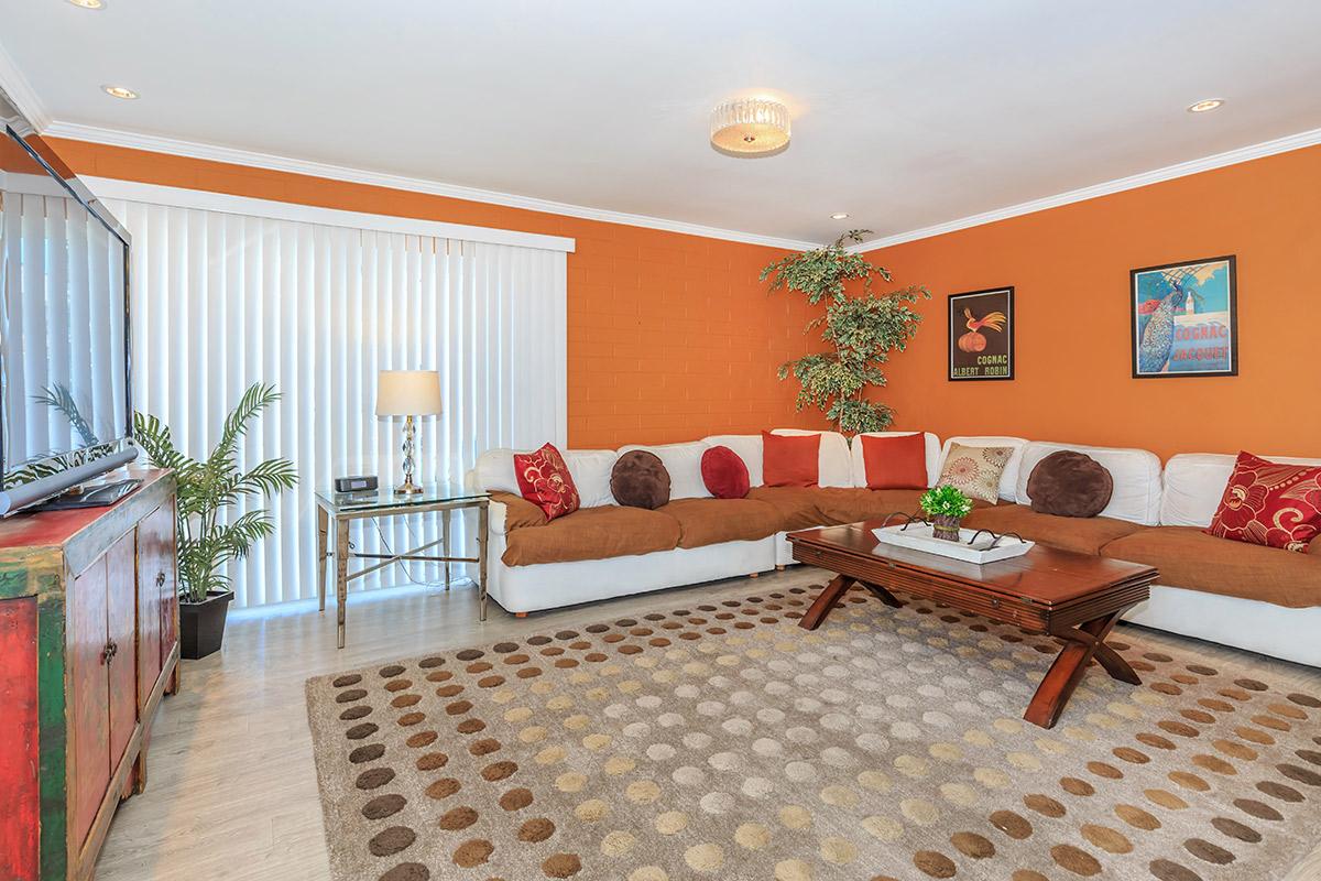 A cozy living room featuring an orange accent wall, a large white sectional sofa with colorful throw pillows, a wooden coffee table, and a patterned rug. There is a television on a stylish cabinet, a lamp beside the sofa, and decorative wall art. A potted plant adds a touch of greenery to the space.