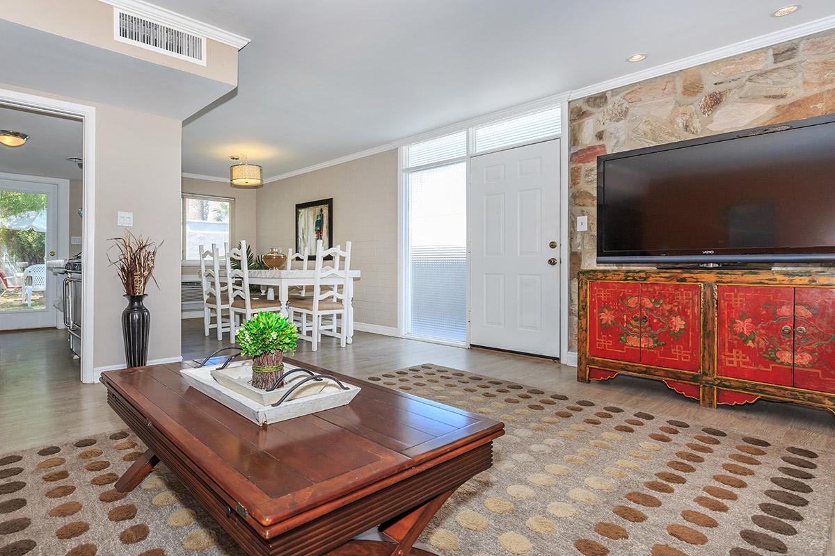 A modern living room featuring a wooden coffee table on a patterned rug, a flat-screen TV mounted on the wall, and a decorative red cabinet. In the background, a bright dining area with a white table and chairs is visible through an open doorway. Large windows allow natural light to fill the space, enhancing the inviting atmosphere.