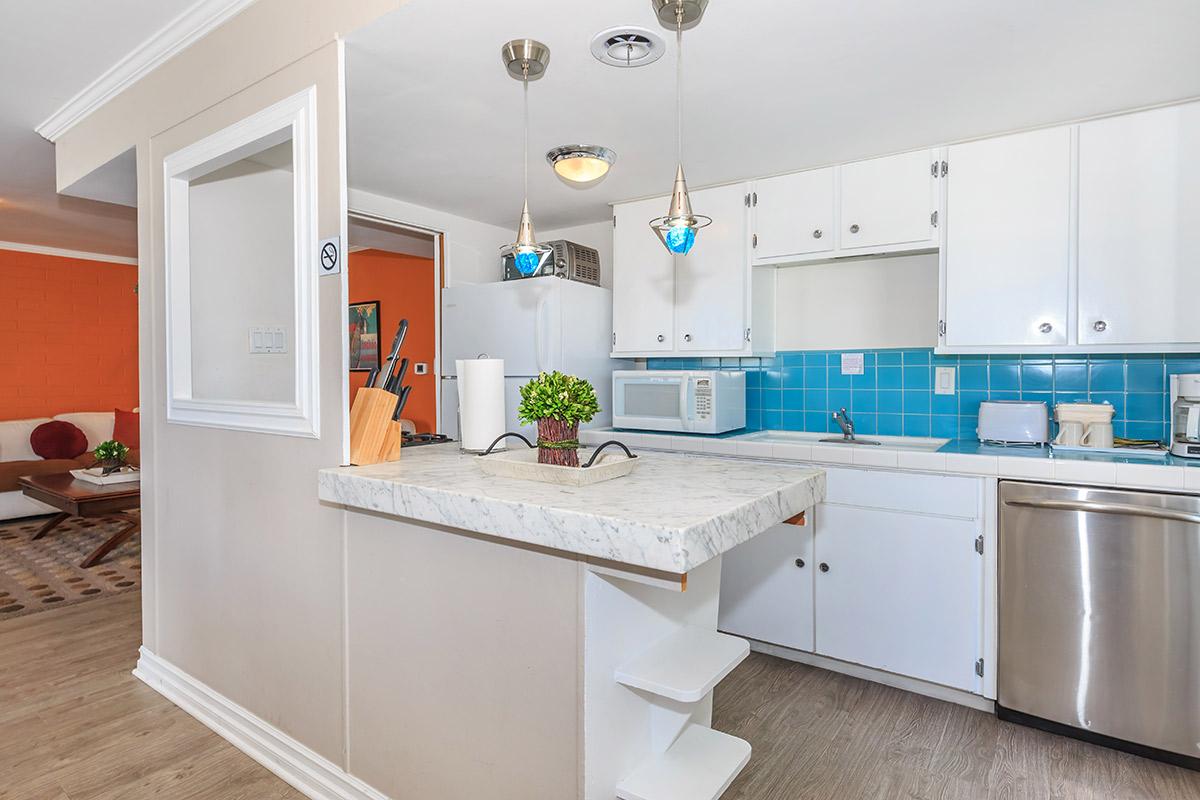 A bright, modern kitchen featuring white cabinetry, a marble countertop, and turquoise tile backsplash. The kitchen is open to a living area, with pendant lights above the bar area and appliances visible, including a refrigerator and microwave.