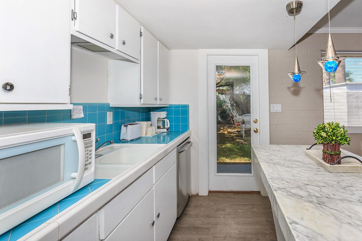 A modern kitchen with white cabinets, a blue tiled backsplash, a microwave, and coffee maker on the counter. There is a marble-top island and a sliding glass door leading outside.