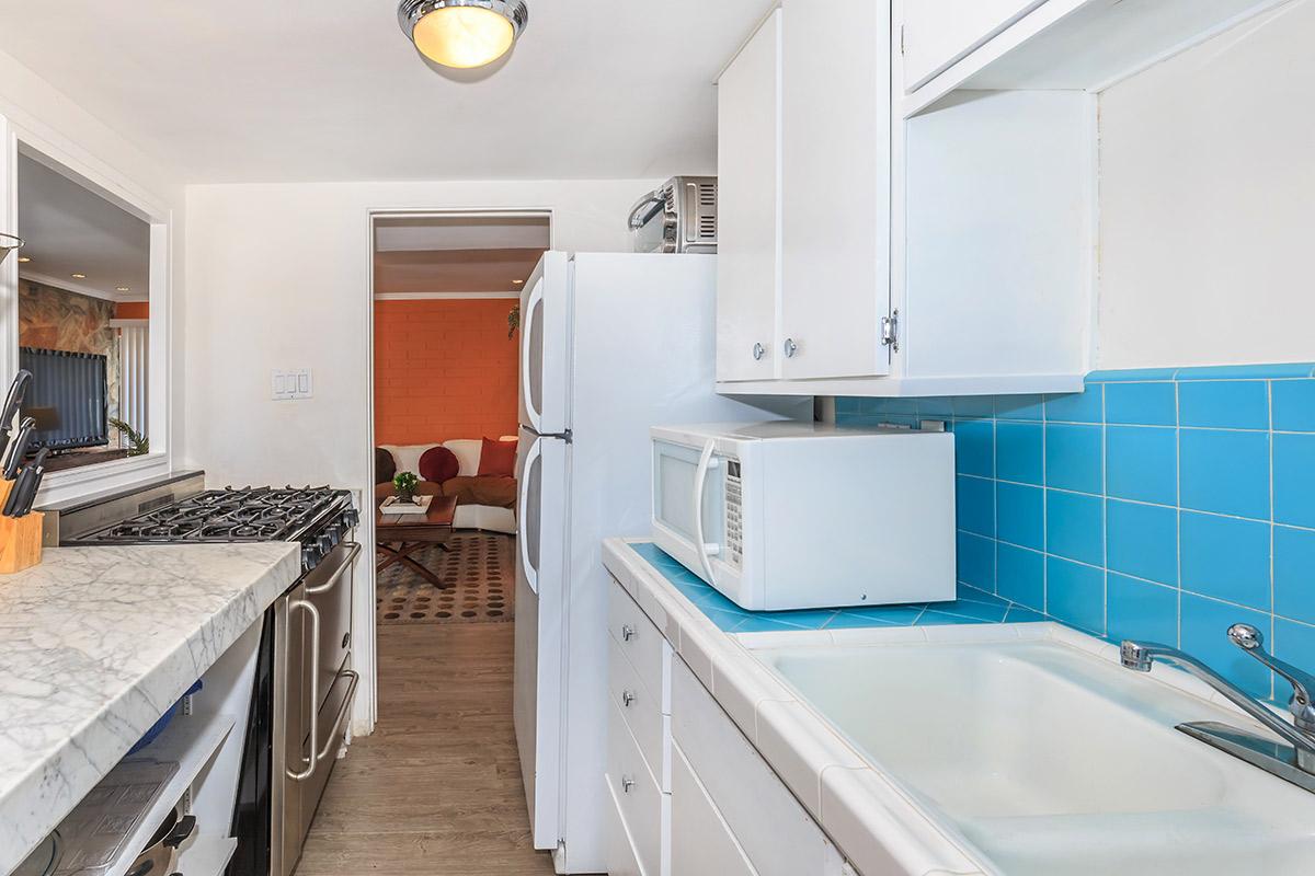 A bright kitchen featuring white cabinets, a marble countertop, a blue tiled backsplash, a stainless steel gas stove, a refrigerator, and a microwave, with a view into a living area beyond.