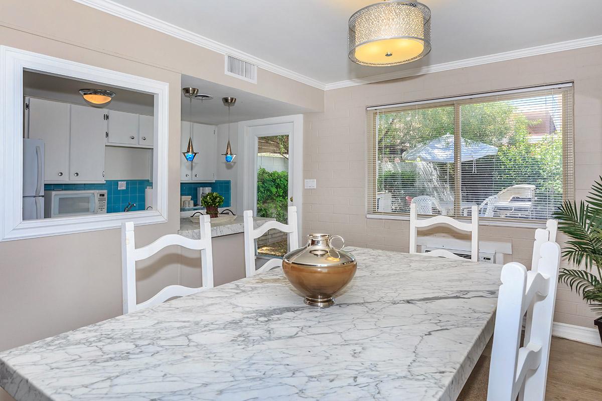 A dining area featuring a large marble table with a decorative vase on it. The room has white chairs and a modern light fixture. A window displays a view of a patio with outdoor furniture and greenery.