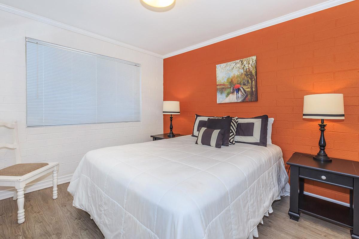 A cozy bedroom featuring a queen-sized bed with white linens, two black nightstands with lamps, and an orange accent wall adorned with a painting. There is a window with blinds allowing natural light, and a small chair beside the bed. The floor is laid with wood-like flooring.