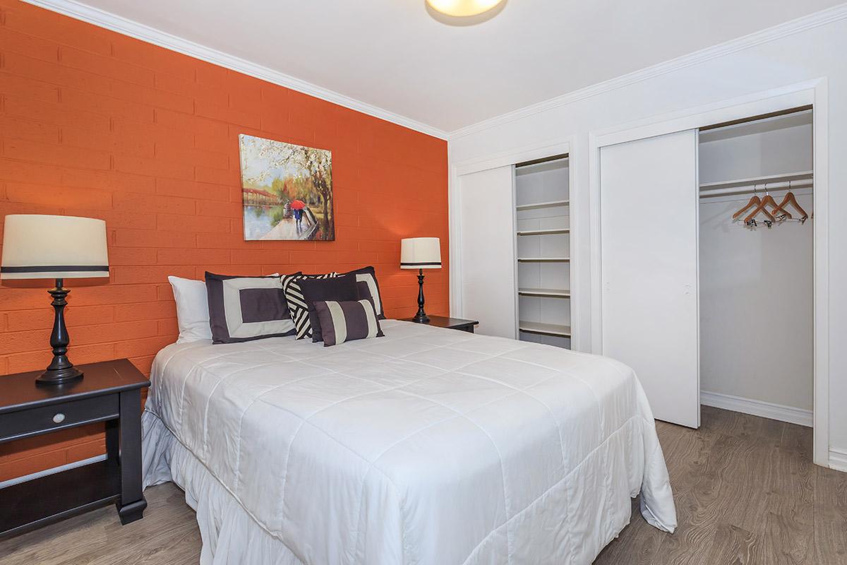 A cozy bedroom featuring a queen-sized bed with white bedding and decorative pillows, two bedside tables with lamps, an orange accent wall with a painting, and a closet with sliding doors.