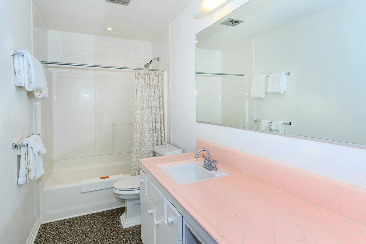 A clean and bright bathroom featuring a bathtub with a shower curtain, a toilet, and a sink with a pink countertop. Towels are neatly hung on racks, and there is a large mirror above the sink. The floor is tiled in dark colors, contrasting with the light walls and fixtures.