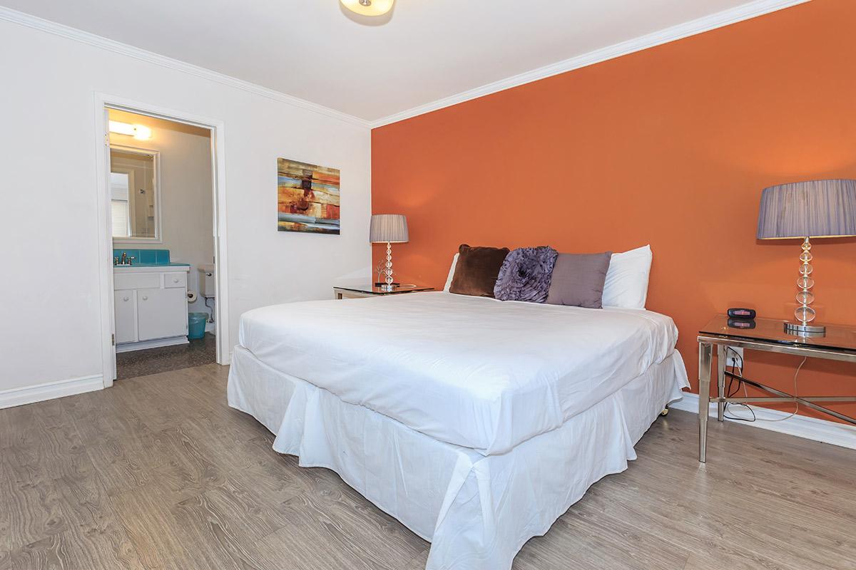A cozy bedroom featuring a large bed with white bedding, two stylish bedside lamps on silver stands, and an orange accent wall. A doorway leads to a bathroom, and artwork is displayed on one wall. The floor is a light wood finish.