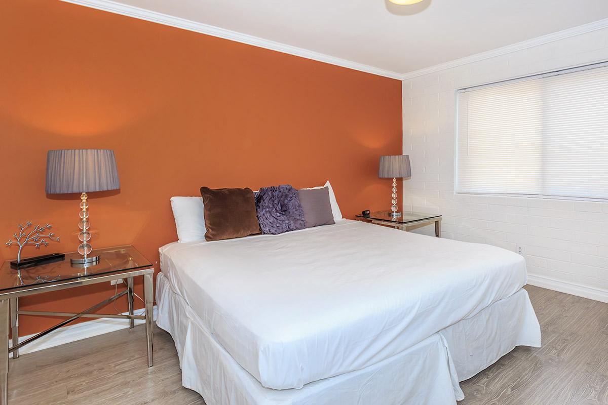 A cozy bedroom featuring a white bed with decorative pillows, two bedside tables with lamps, and an orange accent wall. Natural light filters in through a window with white blinds.