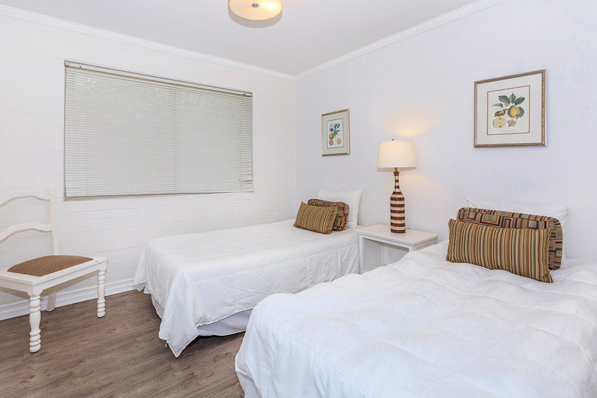A bright and tidy bedroom featuring two twin beds with white bedding and decorative pillows. There’s a small bedside table with a lamp, a chair against the wall, and framed botanical prints on the walls. A window with blinds allows natural light to enter the room.