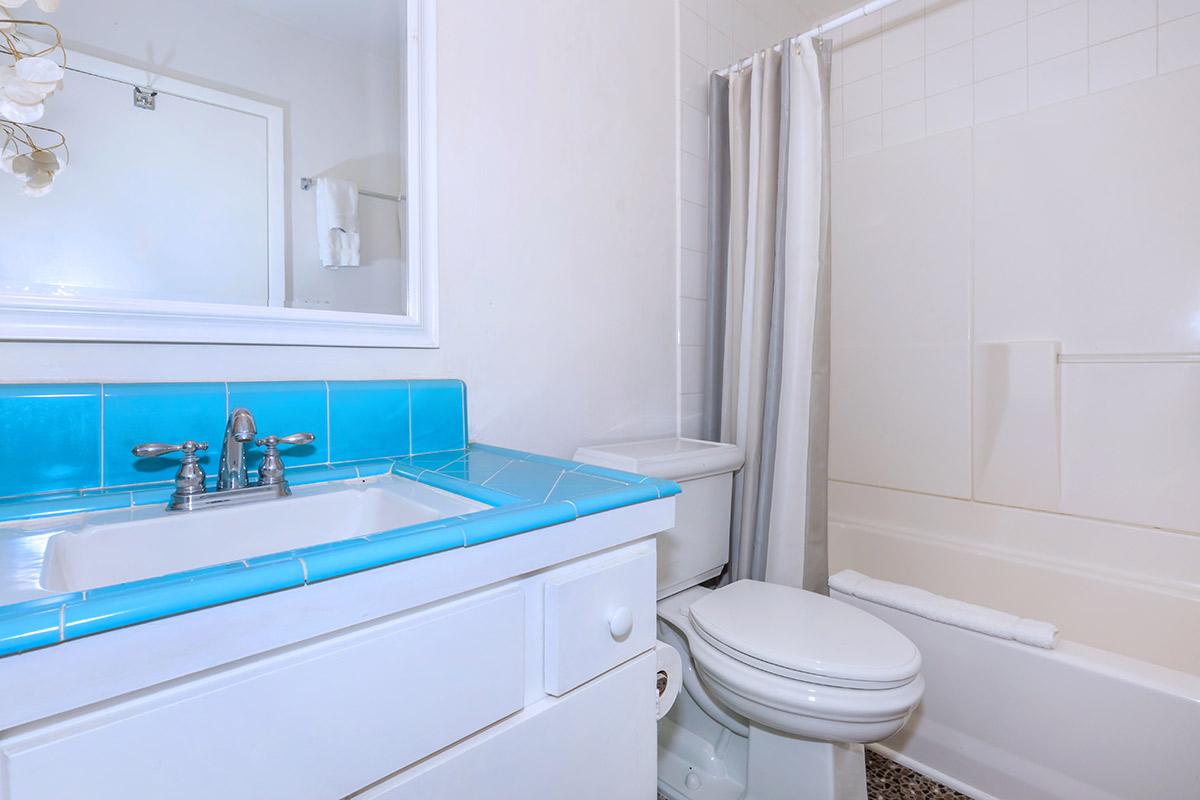 Brightly lit bathroom featuring a blue-tiled countertop with a sink and faucet, a white toilet, and a bathtub with a shower curtain. There is a mirror above the sink and a towel rack on the wall.