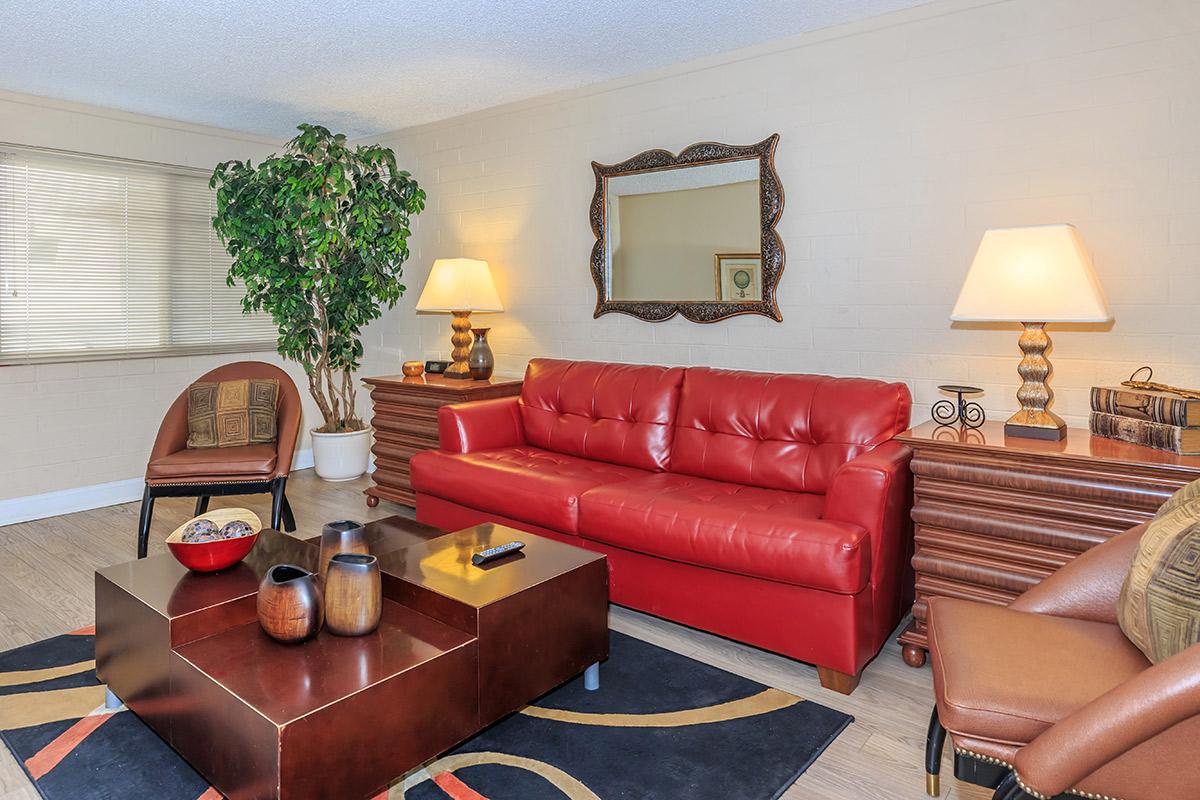 A cozy living room with a red leather sofa, two decorative lamps on wooden side tables, a coffee table with decorative bowls and vases, and a large green plant in the corner. A mirror hangs on the wall above the sofa, and the flooring is wooden with a patterned area rug.