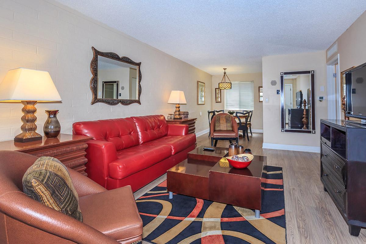 A cozy living room featuring a red leather sofa, a coffee table with decorative items, and a warm-toned armchair. There's a dining area in the background with a wooden table and chairs, and wall mirrors reflect the light, enhancing the space's brightness.