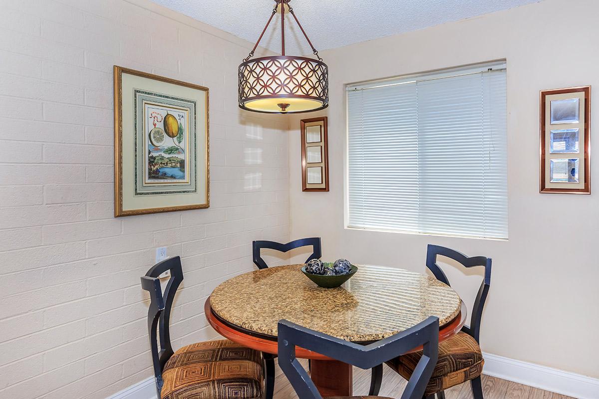A cozy dining area featuring a round table with a granite top, surrounded by three upholstered chairs. A decorative lamp hangs above the table, and a framed artwork is displayed on the wall. There is a window with blinds, allowing natural light to enter the space.