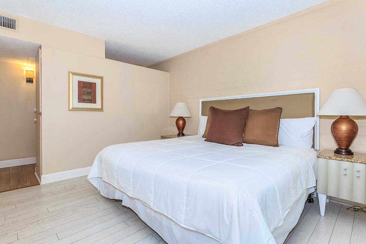 A well-lit bedroom featuring a large bed with two brown decorative pillows, bedside tables with brown lamps, and neutral-colored walls.