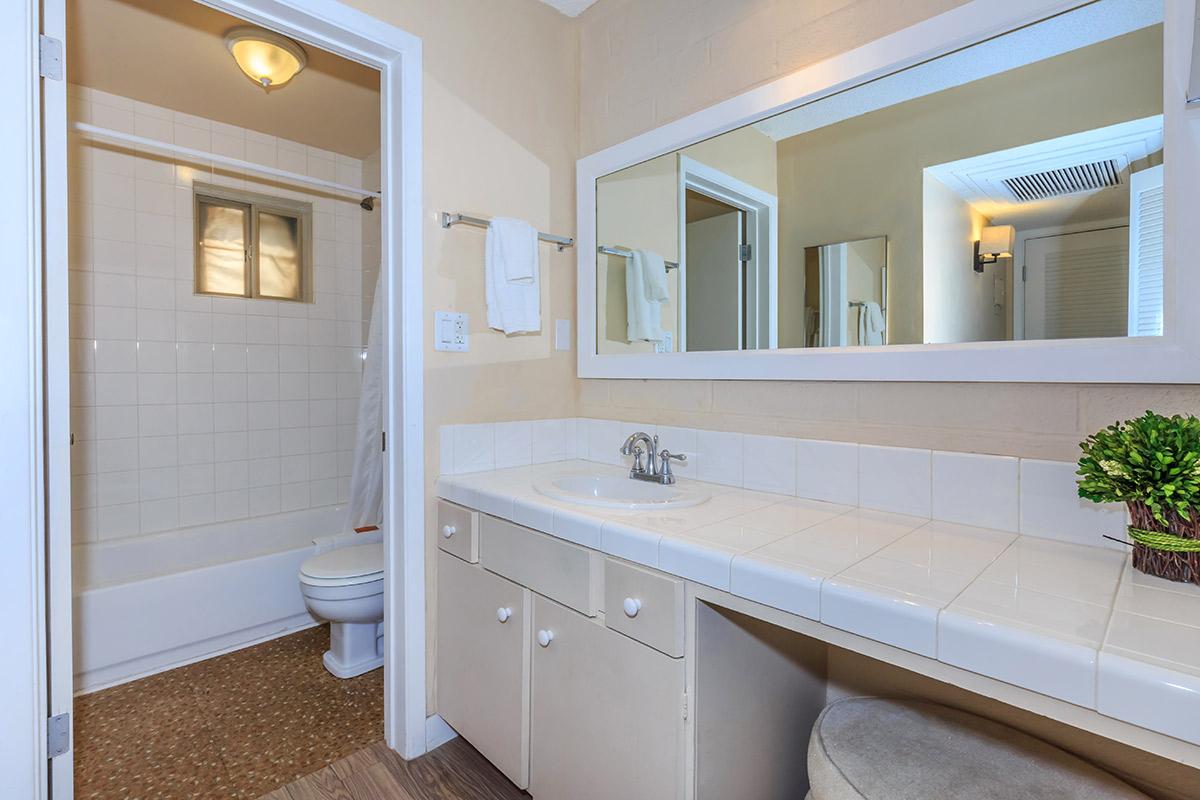 A clean, bright bathroom featuring a white tiled counter with a sink, a mirror, and a small potted plant. A toilet and a shower with a curtain are visible in the background. The walls are painted a light color, and there is a towel rack on the side.