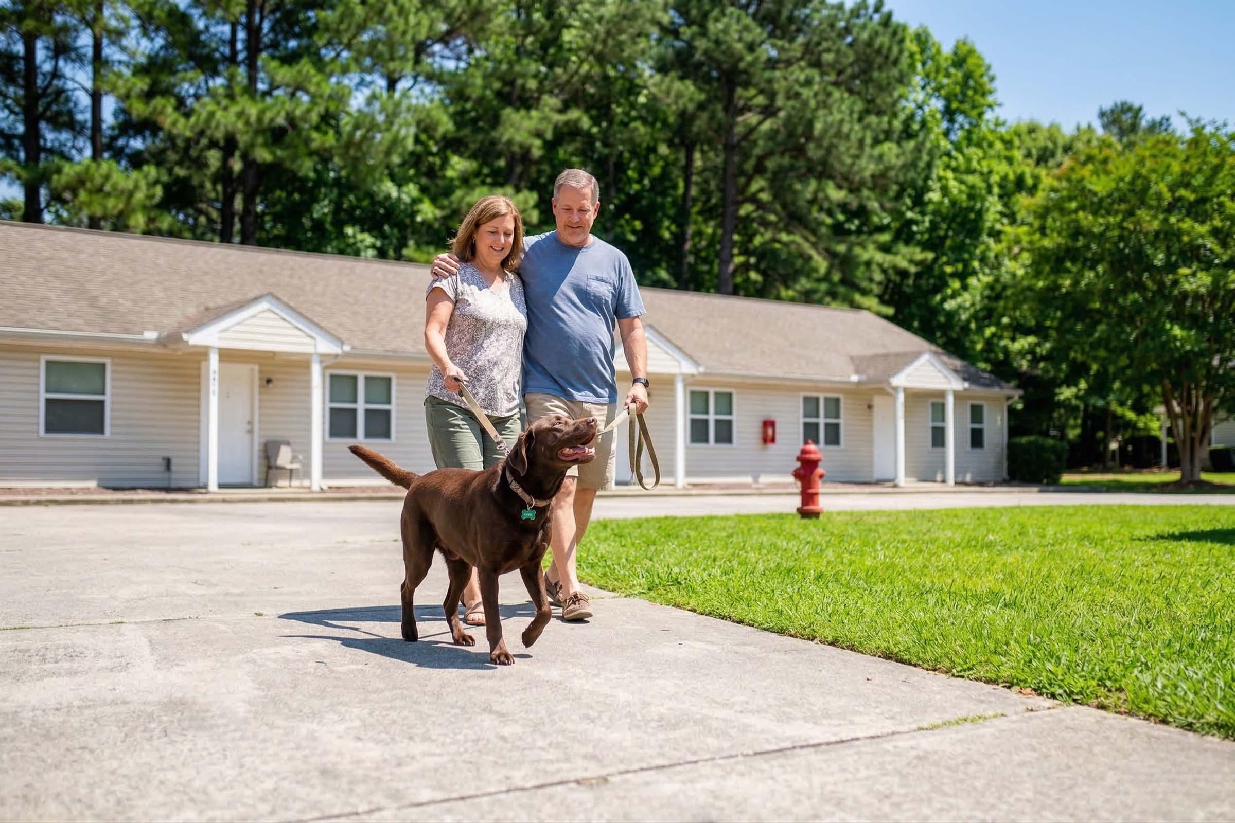 A woman and a man walk a brown Labrador dog on a leash along a sidewalk. They are in a sunny outdoor setting, with green grass and trees in the background. Simple single-story houses can be seen in the distance. The scene conveys a relaxed outdoor atmosphere.