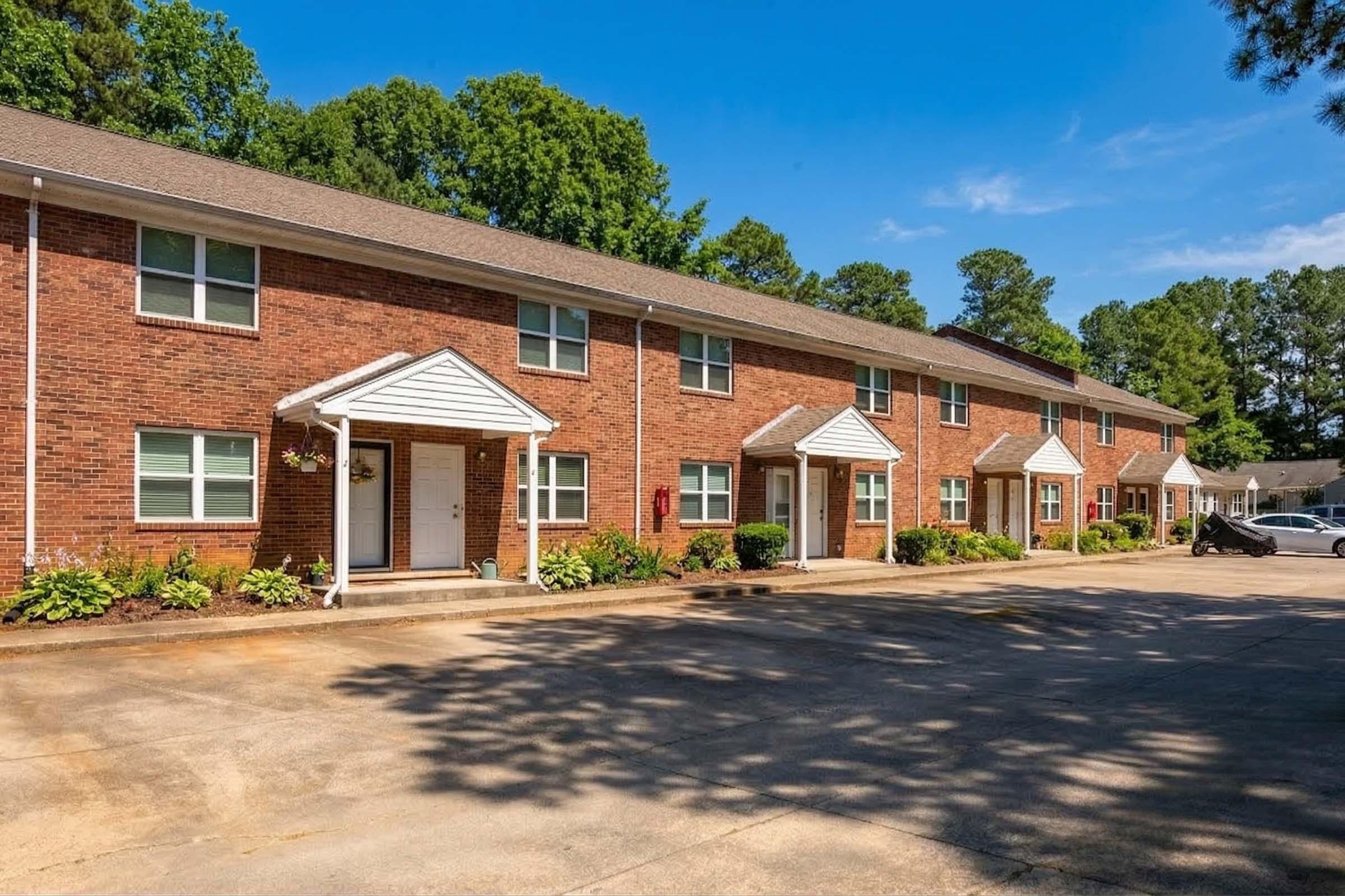 Row of brick apartment buildings with white doors and awnings, surrounded by greenery. A paved pathway leads to the entrances, with flower beds and bushes lining the front. The scene is bright and sunny, showcasing a clear blue sky and a peaceful residential environment.