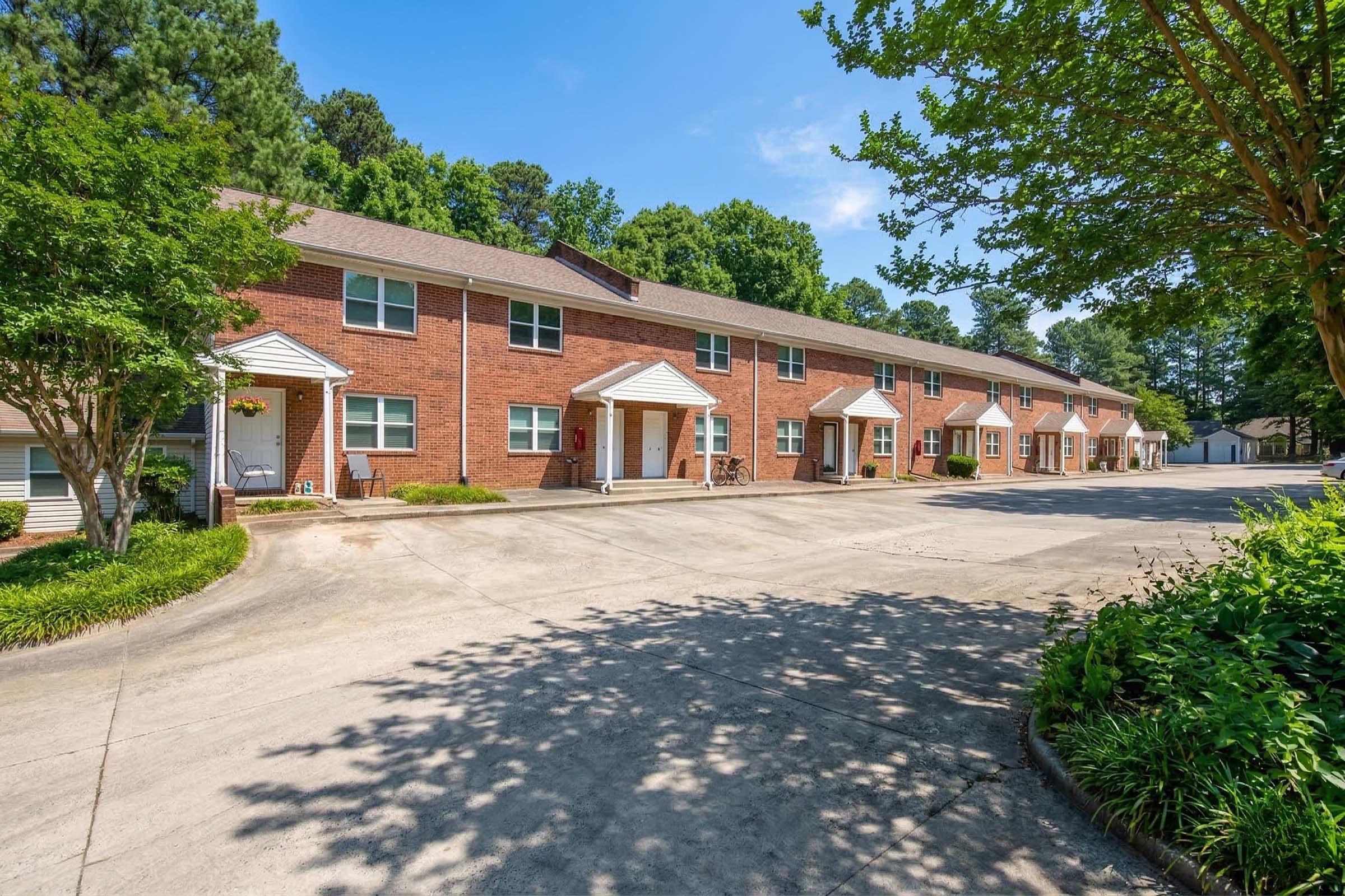 A row of brick apartment buildings with white trim, surrounded by trees and greenery. The pathway leading to the entrances is wide and paved, with a clear blue sky overhead. The setting appears well-maintained and inviting, with a sense of tranquility.