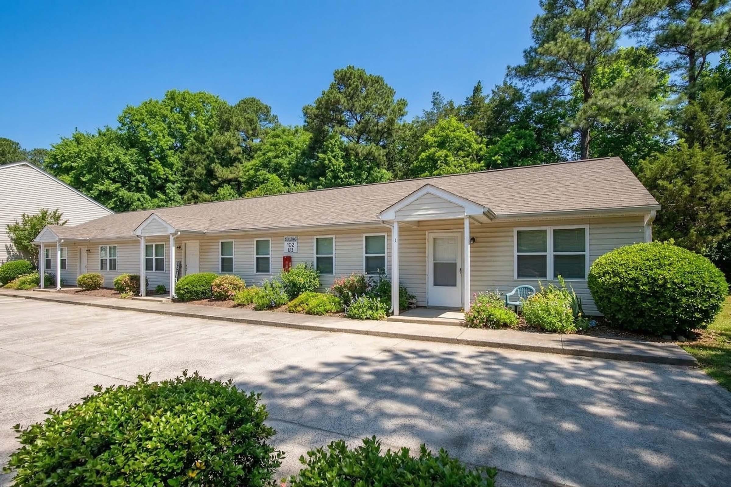 A single-story residential building with multiple units, featuring a light-colored exterior and a sloped roof. The landscaped area includes shrubs and small trees, and there is a paved driveway adjacent to the building. Bright blue sky and greenery in the background create a serene environment.