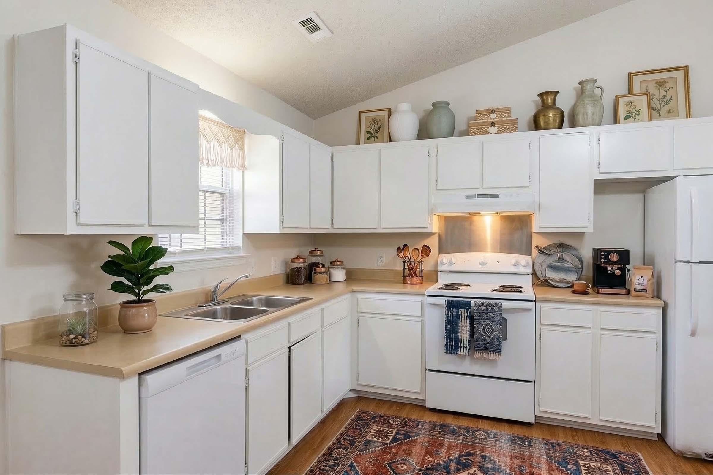 A bright and clean kitchen featuring white cabinets, a beige countertop, and modern appliances. There's a stainless steel sink and a dishwasher. Decor includes a plant on the counter, various kitchen items, and tasteful wall art. A warm light illuminates the space, creating an inviting atmosphere.