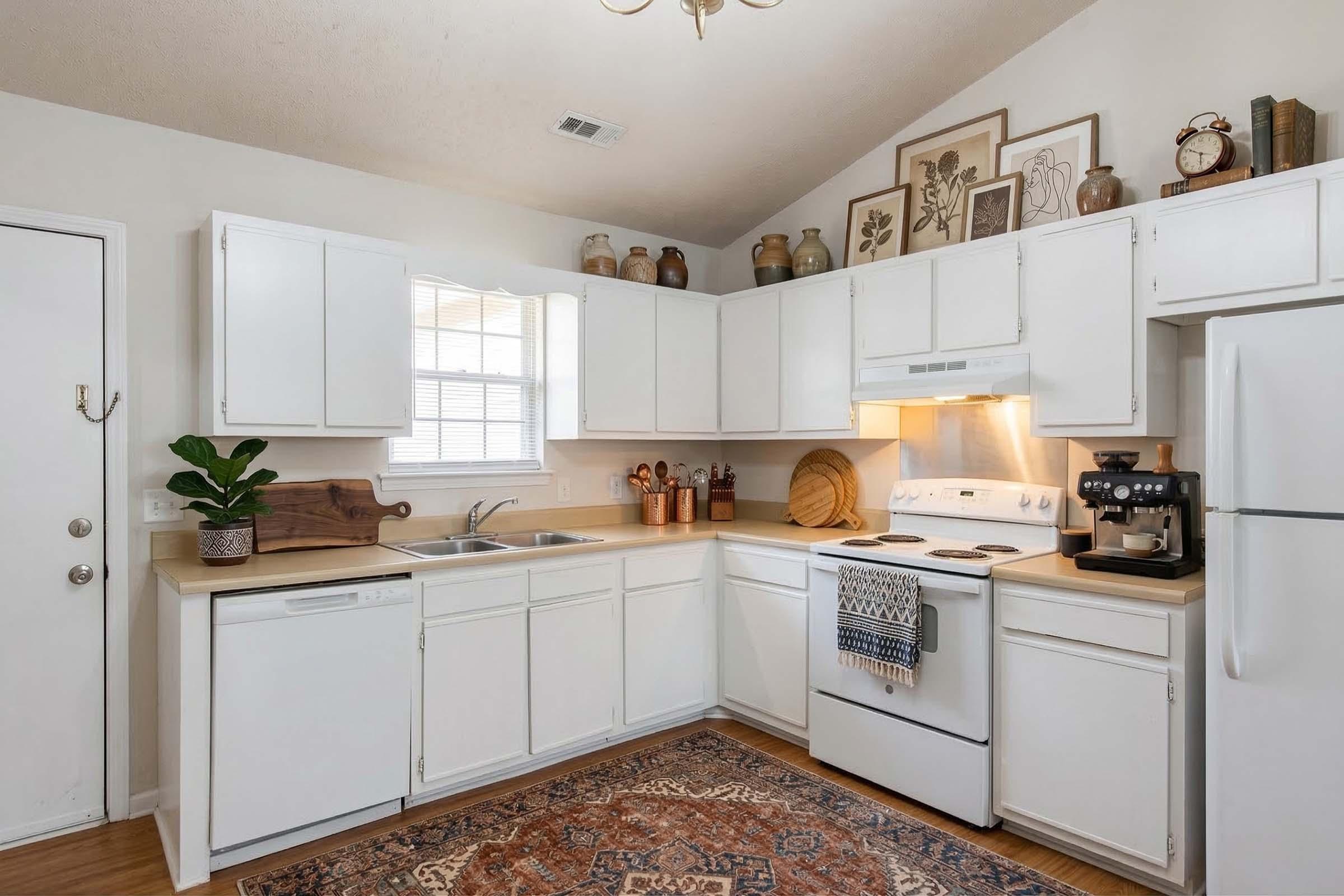 Bright, modern kitchen featuring white cabinetry, a stainless steel sink, and a range with an overhead microwave. There's a coffee maker on the counter, decorative items on shelves, and a patterned rug on the floor, creating a cozy atmosphere. Natural light comes through a window.
