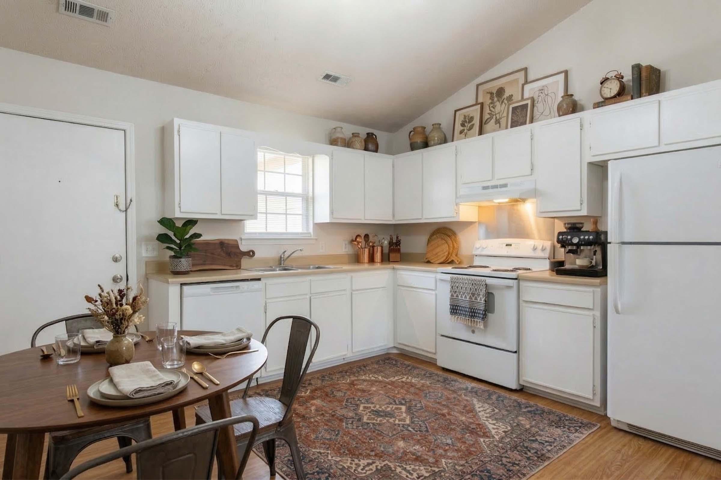A cozy kitchen with white cabinets and wooden countertops. The space features a small round dining table set for two, adorned with plates and utensils. Decorative items and plants are on the shelves, and a patterned rug is underfoot, adding warmth to the room. Natural light streams through a window.