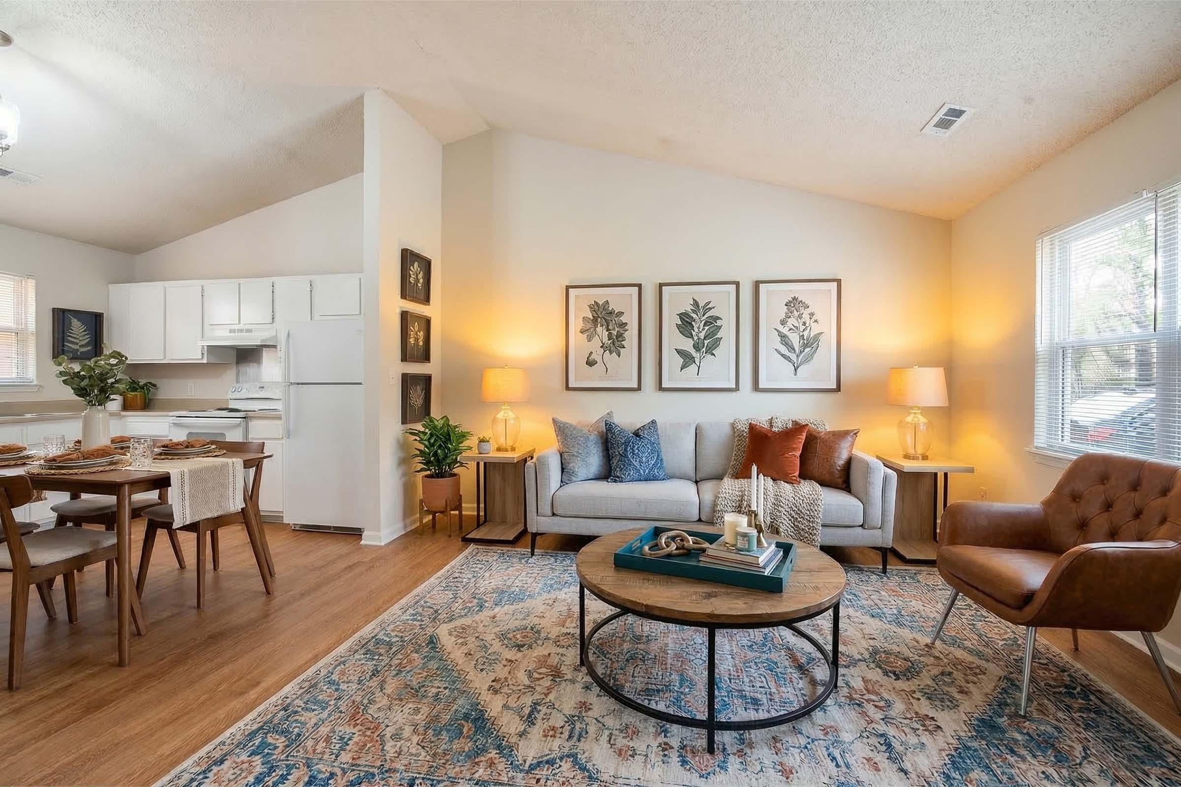 A cozy living room featuring a light gray sofa with decorative pillows, a round coffee table, and an armchair. The walls are adorned with framed botanical prints, and there are lamps on side tables. A dining area with wooden chairs is visible, along with an open kitchen in the background. Natural light streams in from the windows.